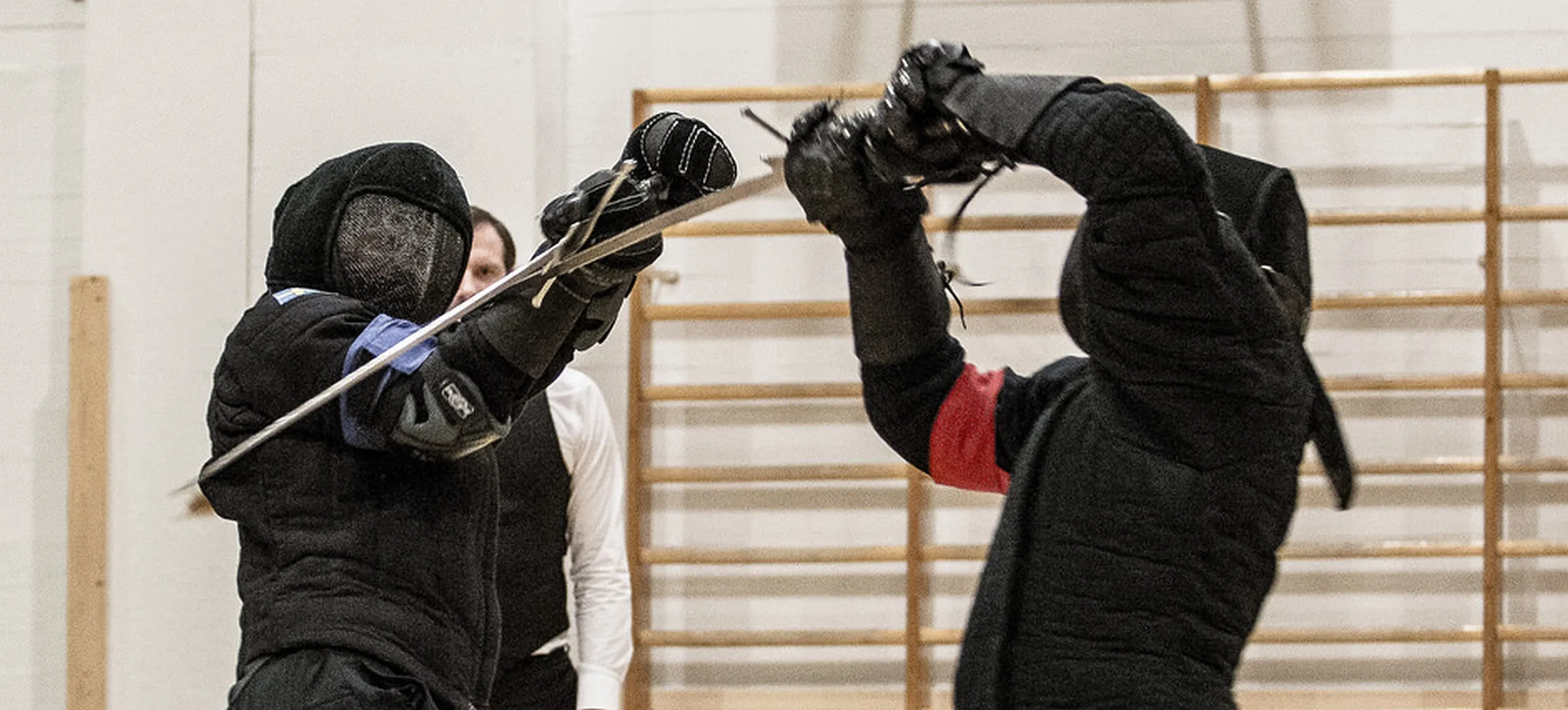 Two fencers in a bind with their swords raised, in the gym hall in front of wall bars.