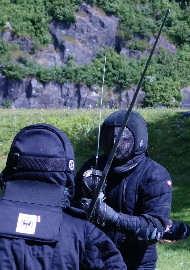 A masked fencer holding a longsword outdoors in a green Norwegian landscape.