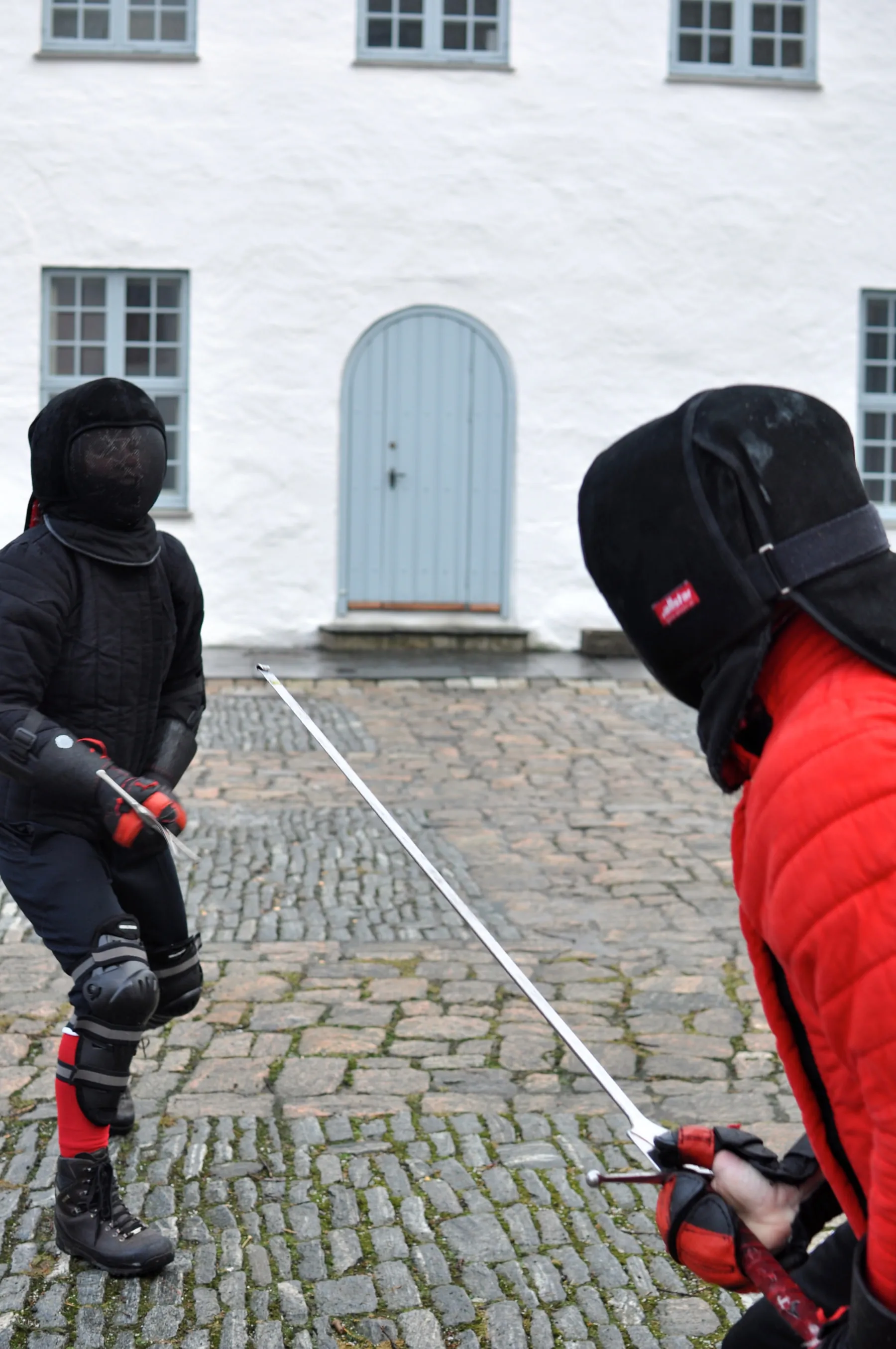 Two fencers in masks and full protection sparring outdoors on cobblestones.
