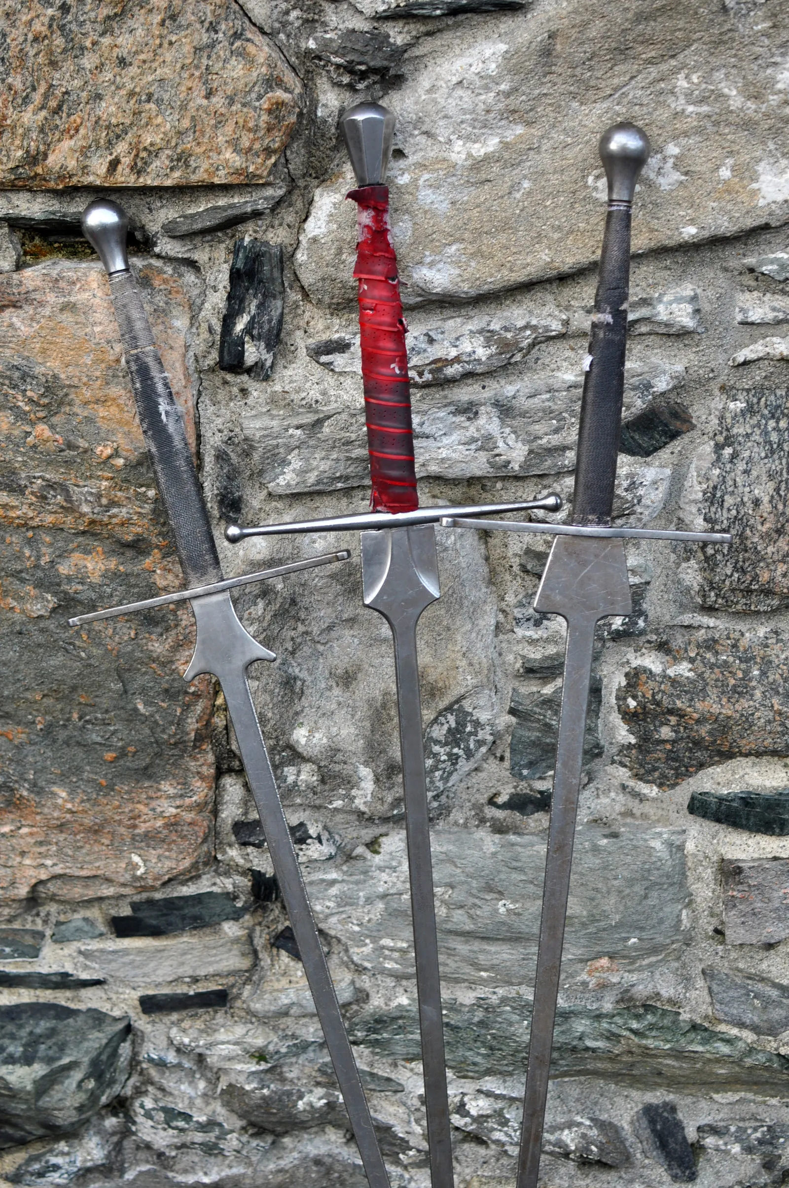 Close-up of a longsword with a red-wrapped grip, leaning against a stone wall.