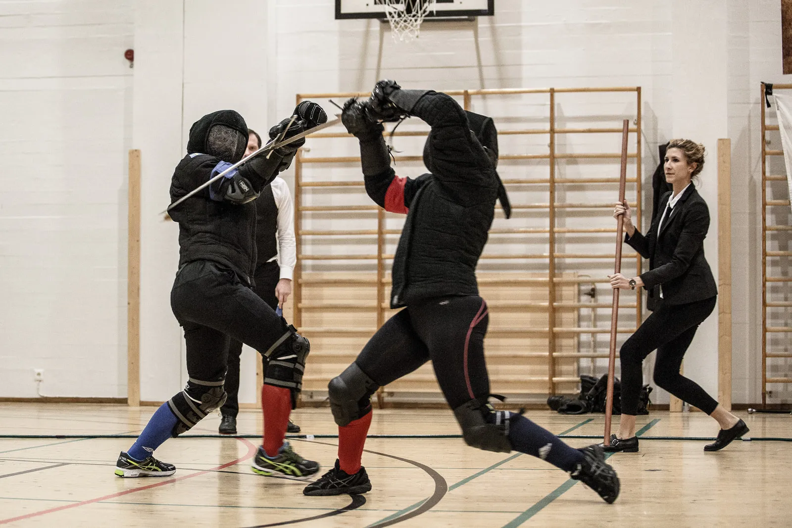 Two fencers sparring in the gym hall, wall bars behind them and swords crossed overhead.